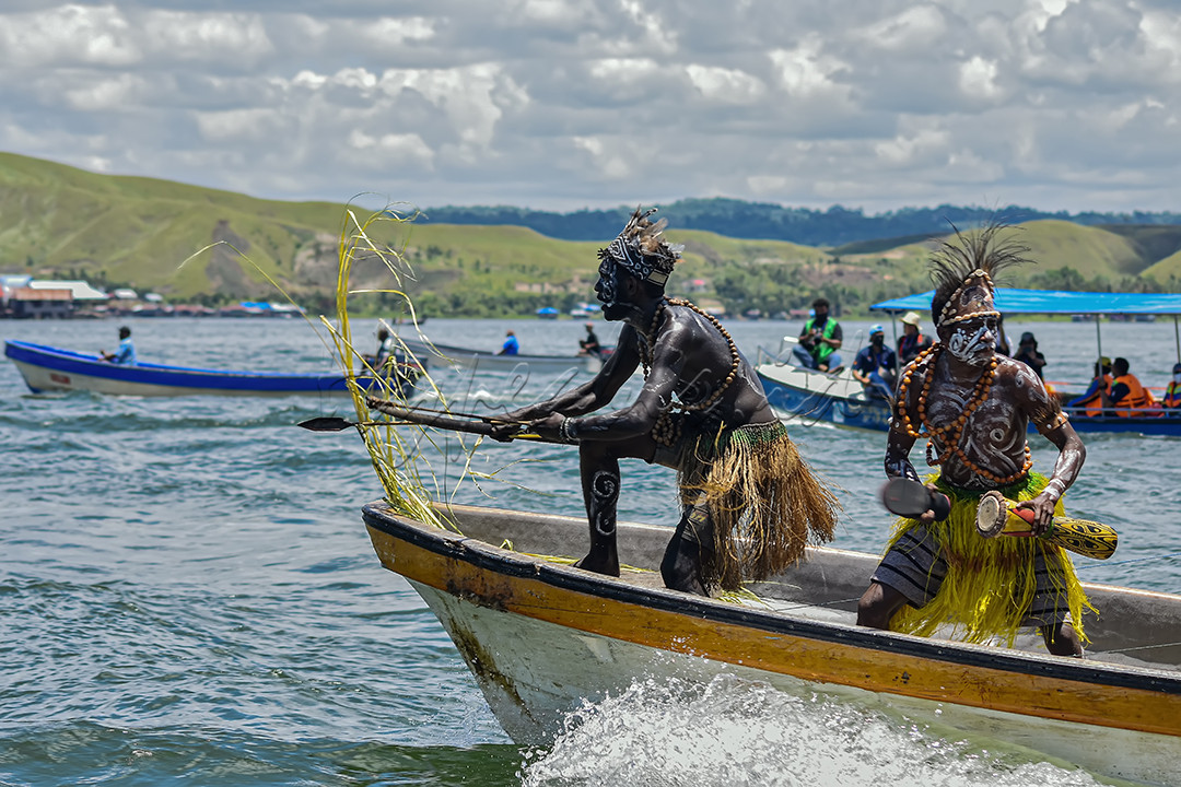 Di dalam foto ini, dapat kita lihat bagaimana kentalnya budaya masyarakat sentani, papua. nampak kedua Pria berpakaian adat itu menari tarian adat mereka diatas speedboat, di tengah-tengah luasnya dan keindahan alam yang asri di danau sentani, Papua.